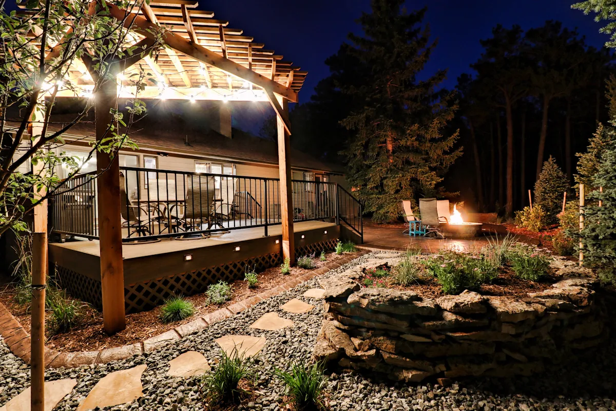 A string-lit cedar pergola over a deck at dusk, with a lit fire pit beyond