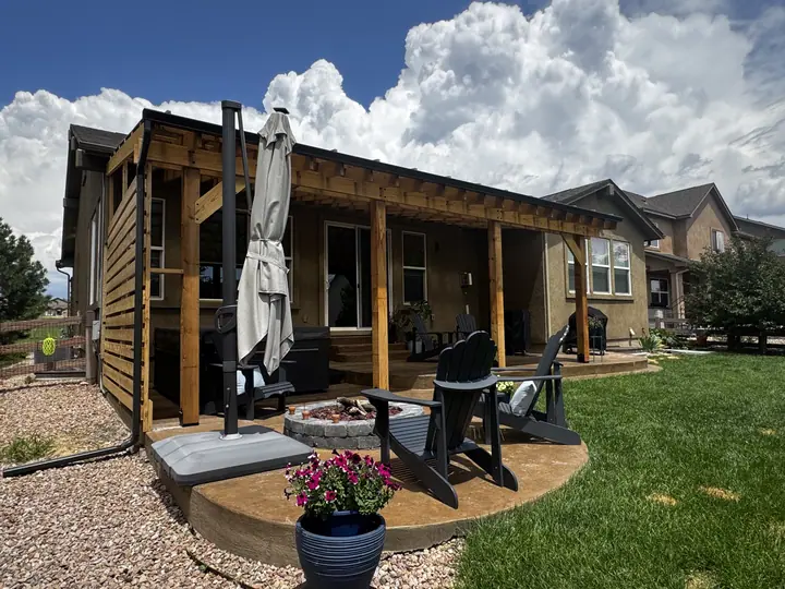 A cedar pergola attached across the back of a house, with a fire pit and chairs on the patio